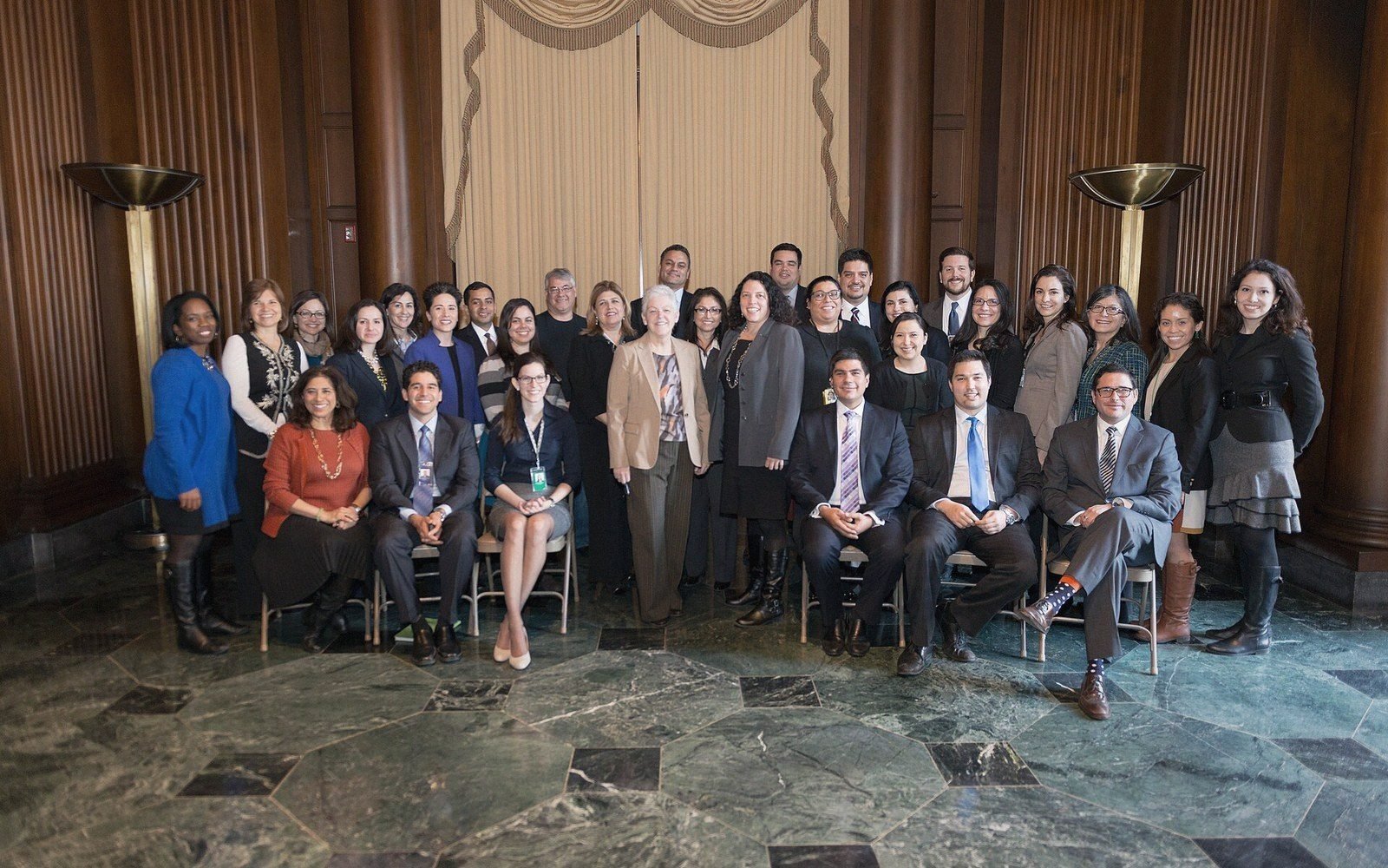 Large group poses together in formal attire on a marble floor in a wood-paneled hall