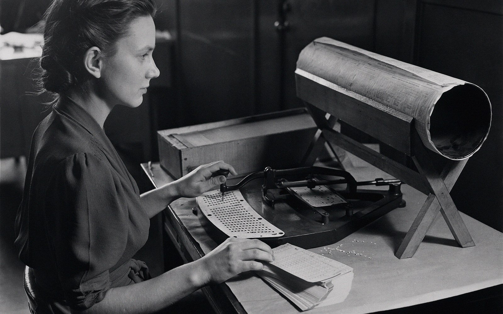 Woman using a mechanical card punch beside a rotating drum reader on a desk