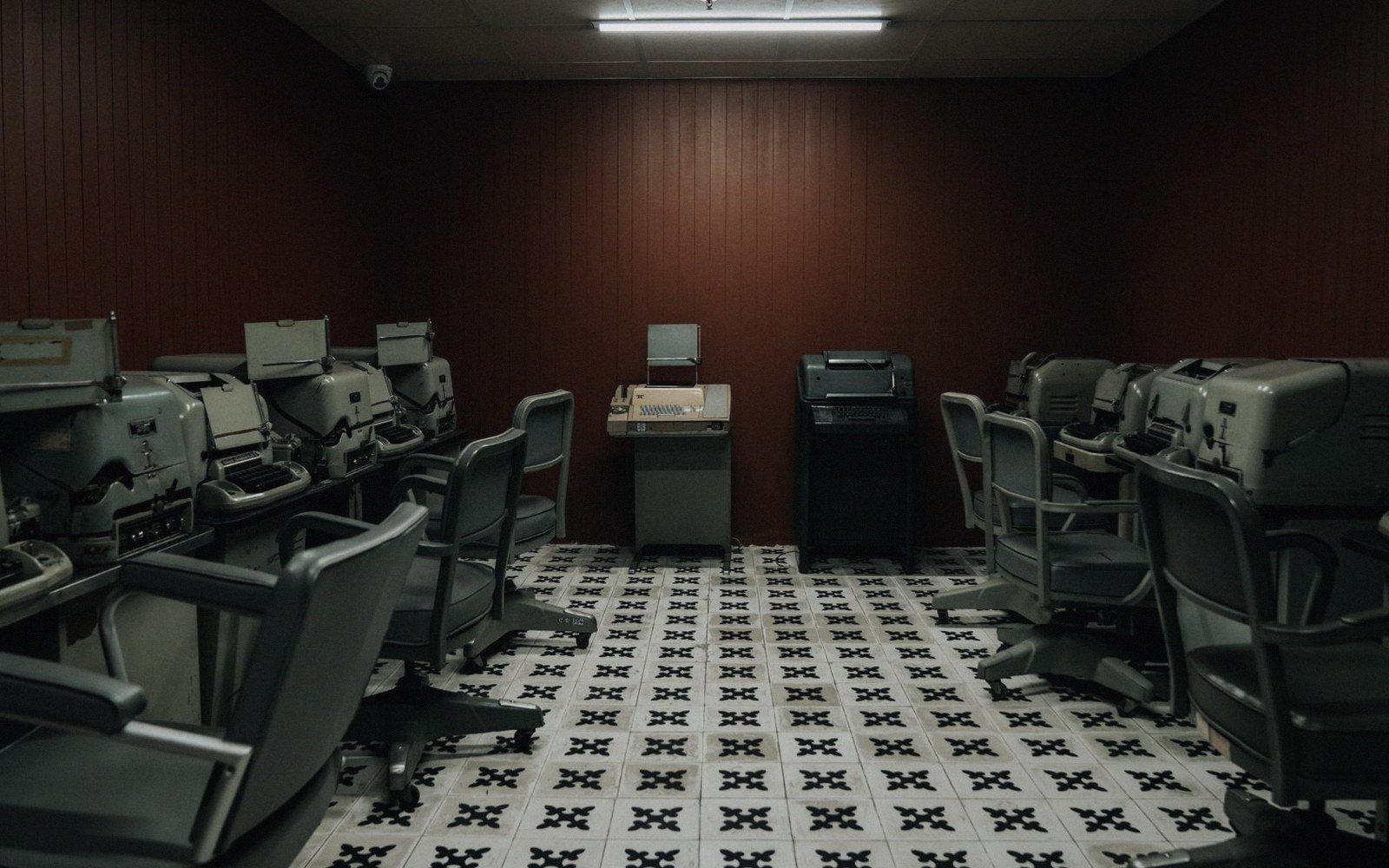 Rows of vintage teleprinter machines and chairs face each other on patterned tile flooring