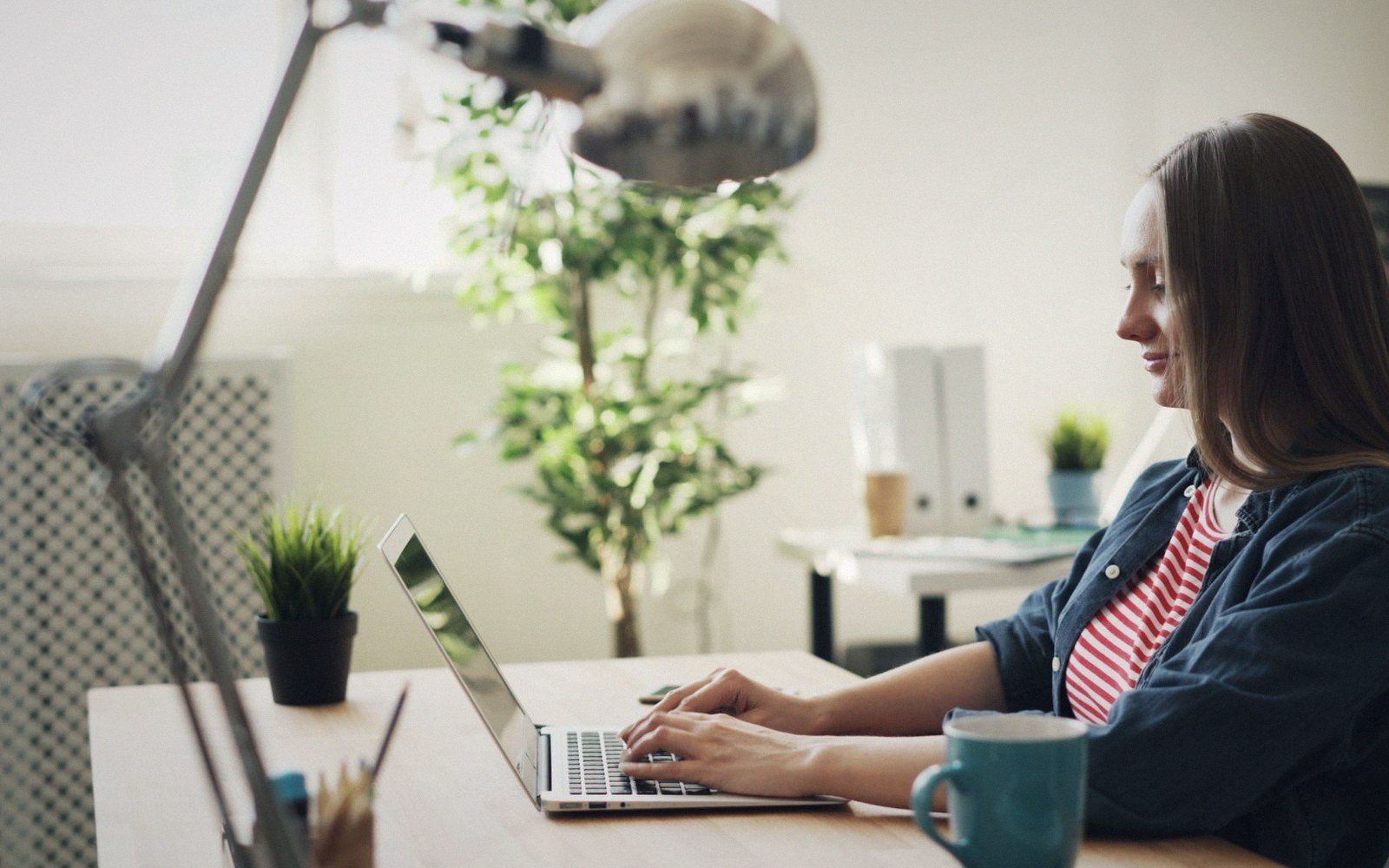 Woman smiling while typing on a laptop at a desk with plants and a mug