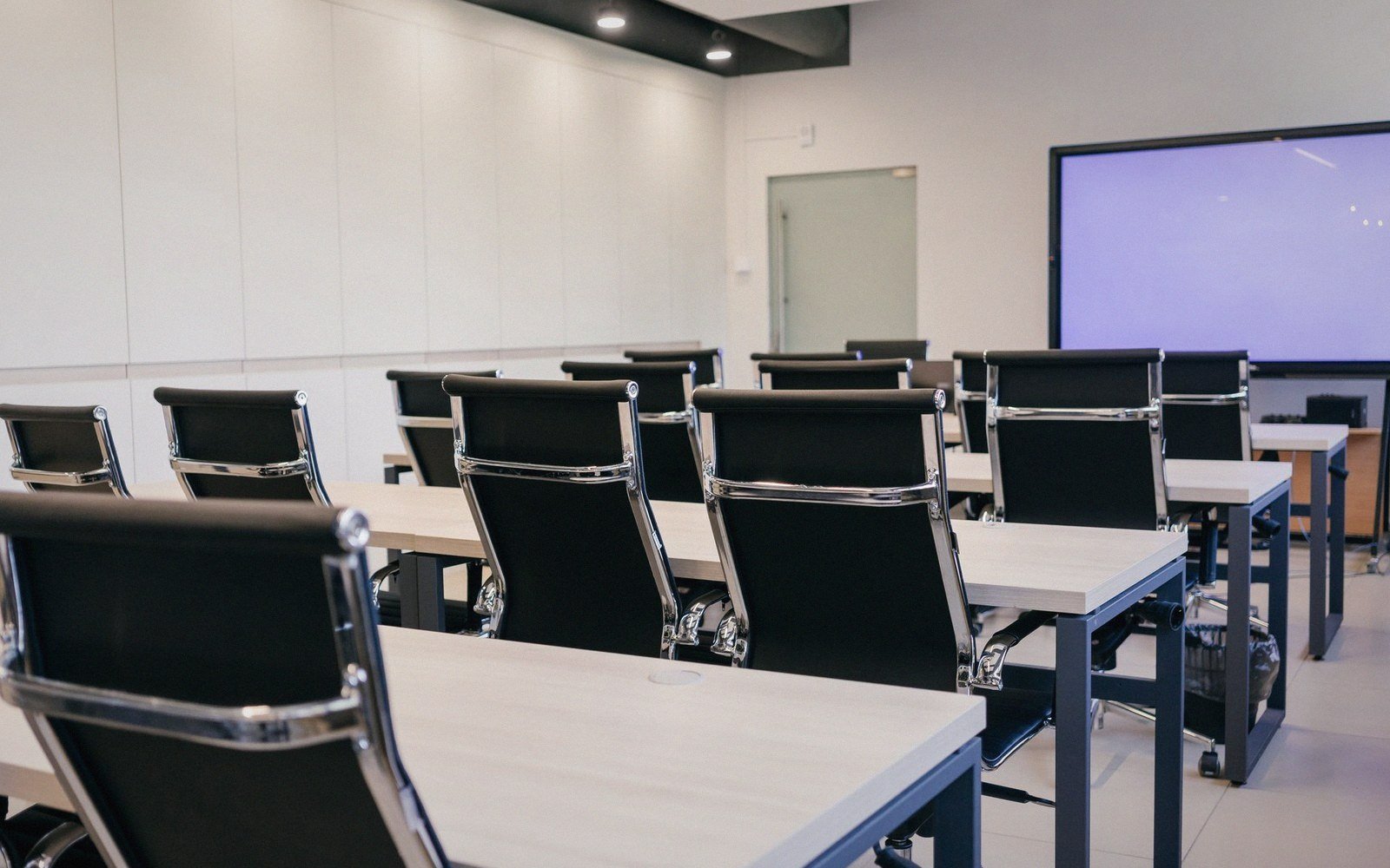 Rows of empty office chairs and tables face a blank screen in a training room